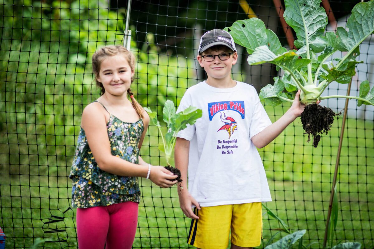 Two children stand in a garden holding leafy greens they have picked. The girl on the left is holding a small plant, while the boy on the right holds a larger one. They are smiling, and a garden net is visible in the background.