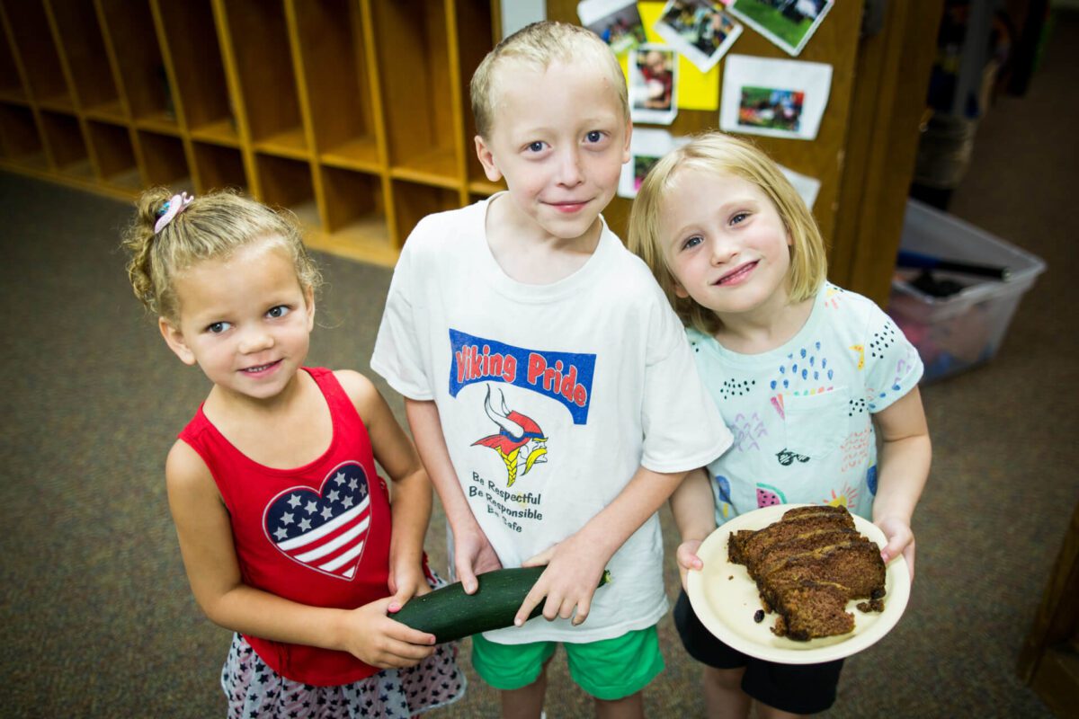 Three young children stand indoors smiling at the camera. The boy in the center holds a large zucchini and wears a shirt with 