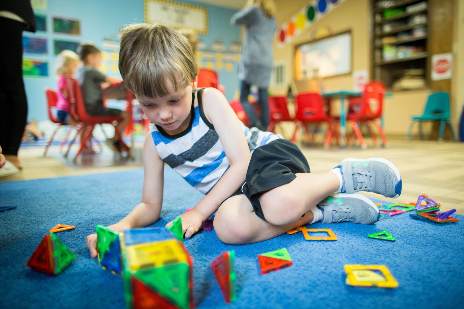 A young child plays with colorful magnetic building blocks on a blue rug in a classroom. Other children and an adult are in the background, surrounded by bright decor and red chairs.