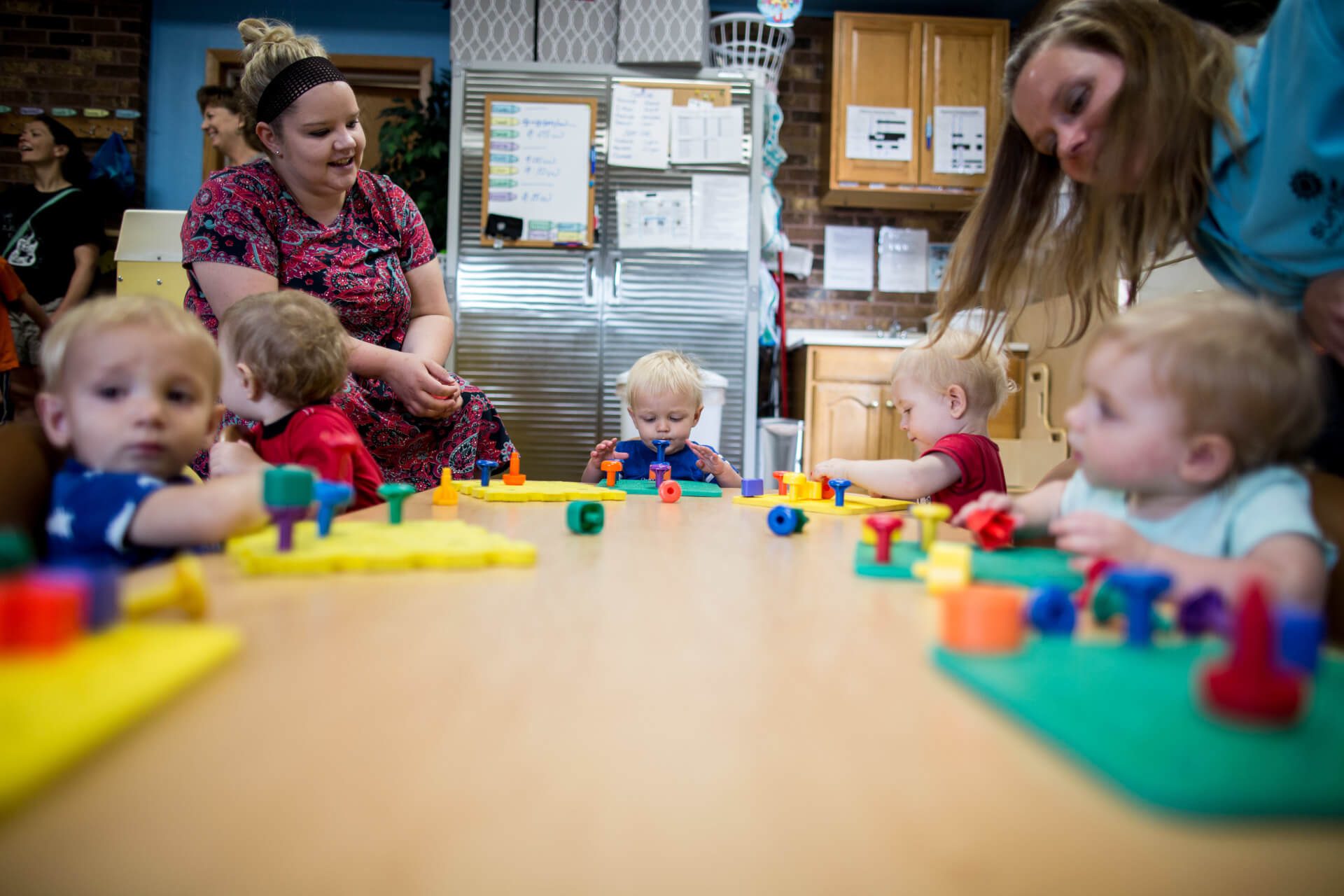 A group of toddlers sit around a table in a daycare, playing with colorful toys. Two caregivers are nearby, watching and engaging with the children. The room has educational posters and a fridge in the background.