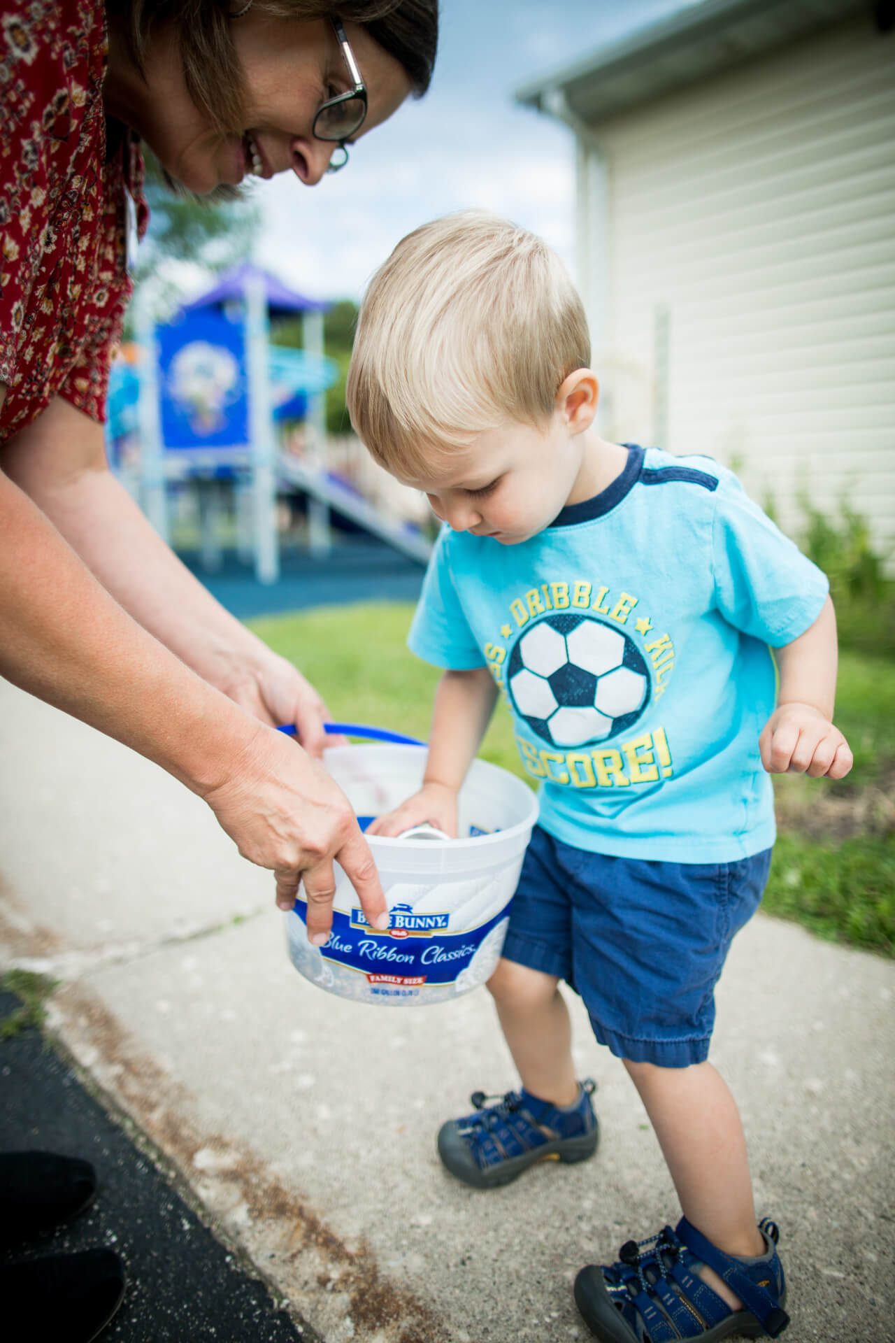 A toddler in a blue shirt and shorts looks into a plastic bucket held by an adult. The adult wears glasses and a patterned red shirt. They stand on a sidewalk near a playground area with blue play structures in the background.