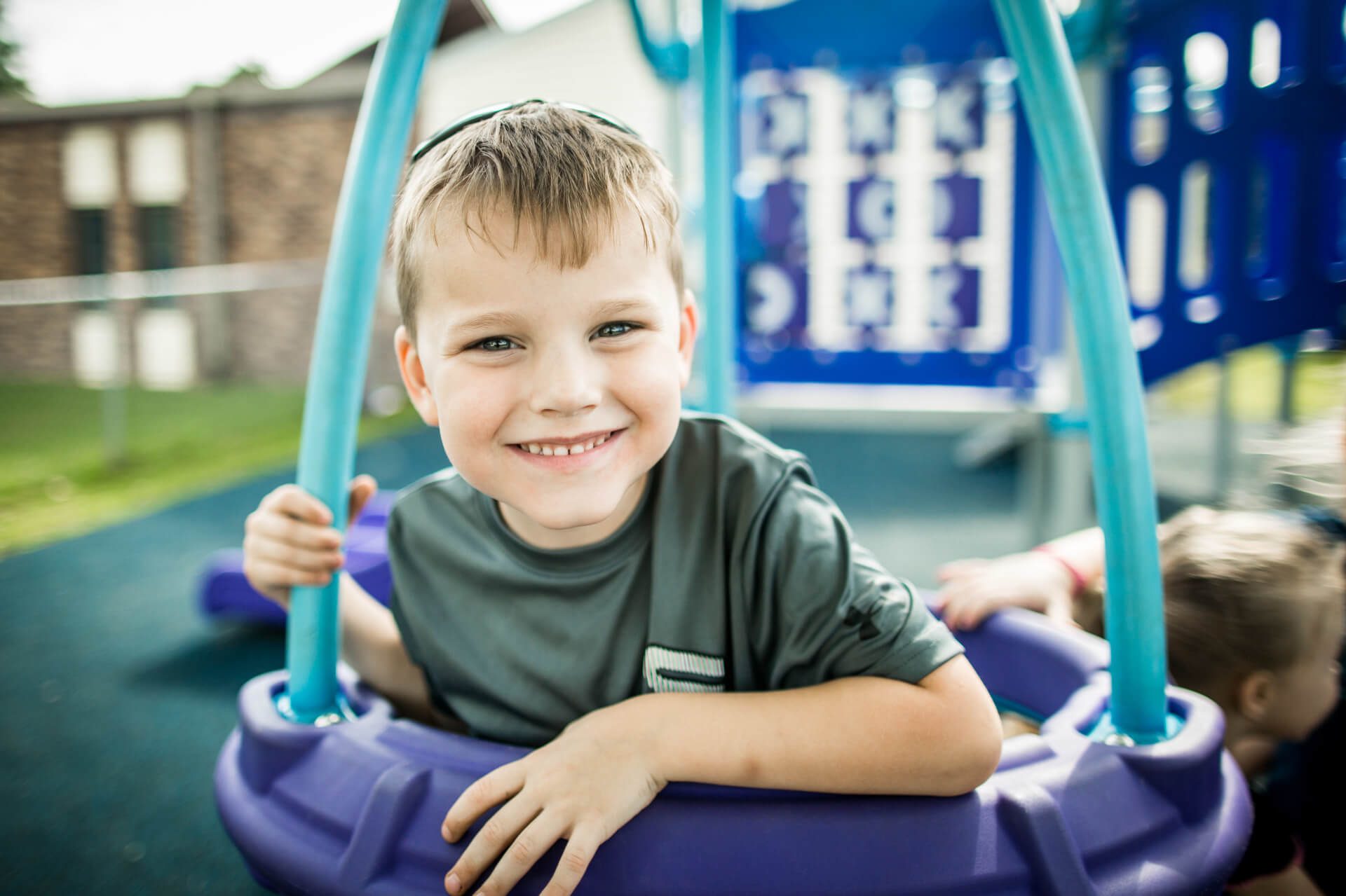 A young boy smiling while holding onto a swing at a playground. He is wearing a gray shirt, and the playground has blue equipment in the background. Another child is partially visible, playing next to him.