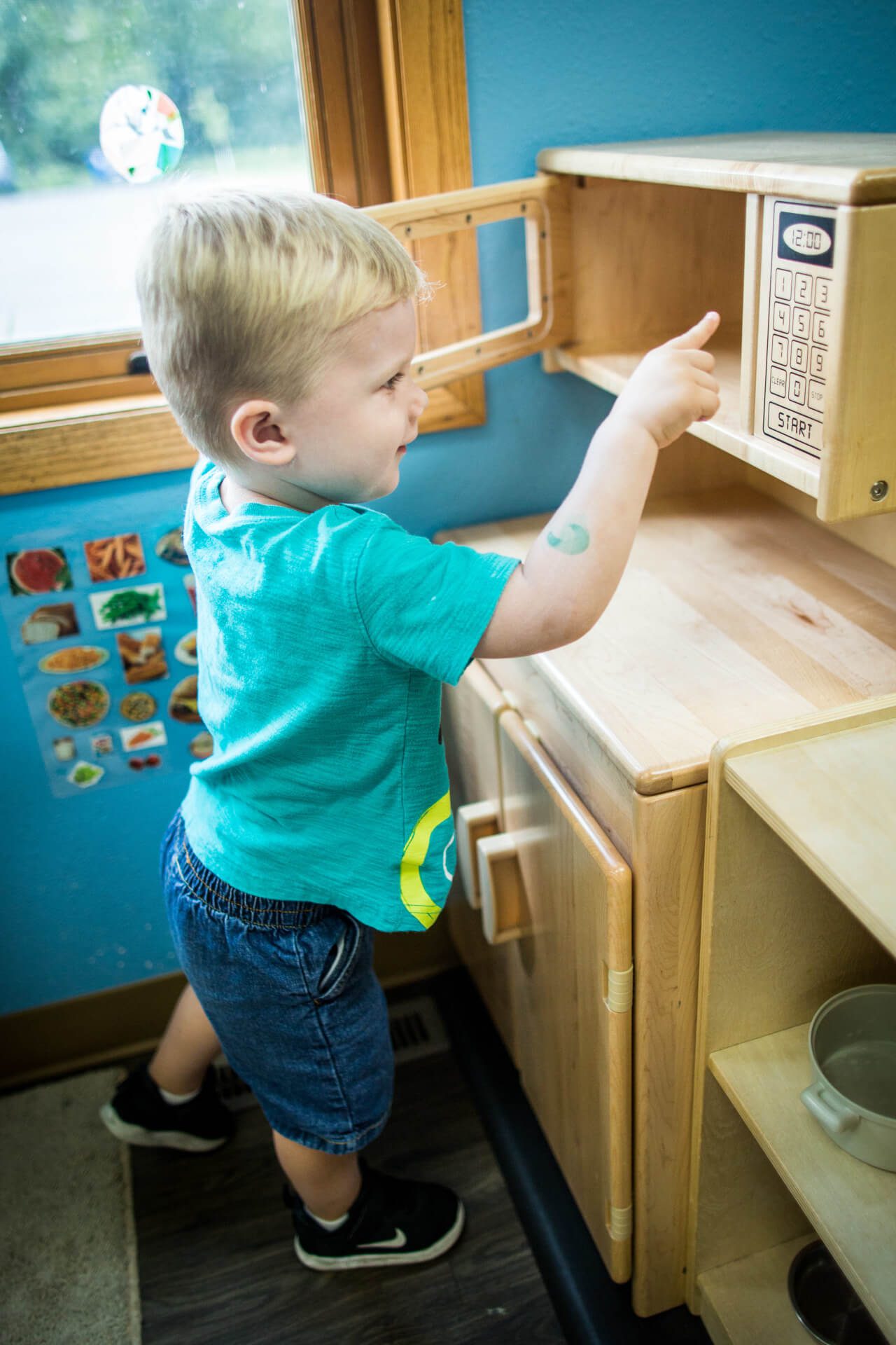 A young child with blonde hair stands in front of a wooden play kitchen set, reaching for buttons on a toy microwave. The child is wearing a turquoise shirt and jean shorts. A colorful food chart is on the wall in the background.