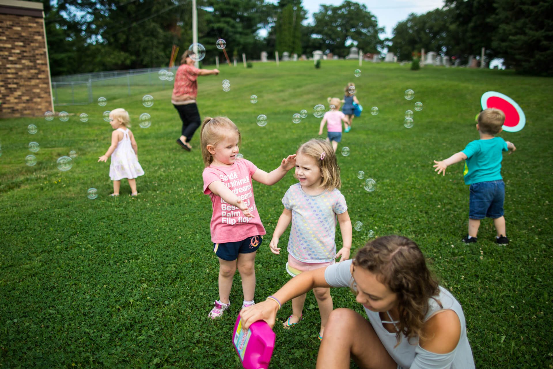 Children play outside on a grassy field, blowing and chasing bubbles. A caregiver pours bubble solution nearby. The scene is joyful and vibrant, with trees in the background on a sunny day.