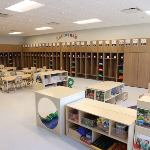 A brightly lit preschool classroom with small tables and chairs, wooden storage cubbies along the walls, and various educational toys and materials. The room is organized and has colorful decorations.