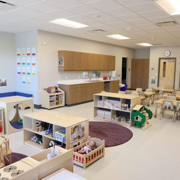 A spacious, well-organized preschool classroom with wooden tables and chairs, shelves filled with toys, books, and educational supplies. The room includes colorful play areas, a kitchenette, and a clean, bright atmosphere with a clock on the wall.