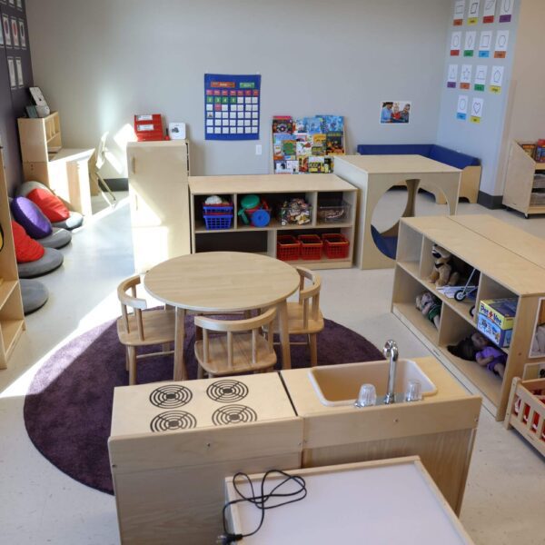 A well-lit preschool classroom with wooden furniture. There's a round table with chairs, shelves stocked with toys and books, a play kitchen set, and colorful cushions. Educational posters decorate the walls.