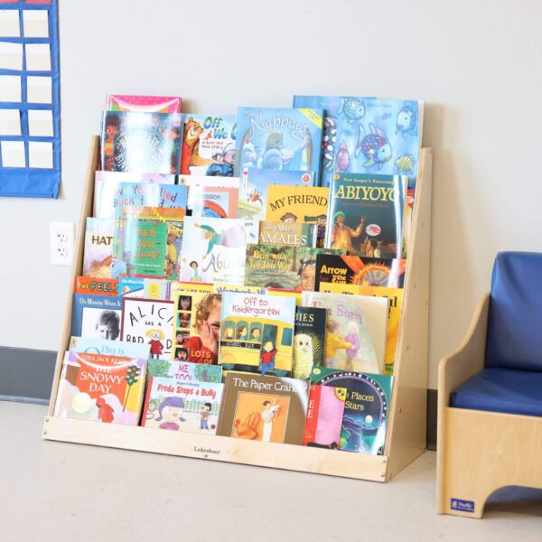 A bookshelf filled with colorful children's books stands against a wall. Nearby, there’s a blue cushioned bench. A colorful calendar is partially visible on the wall to the left.