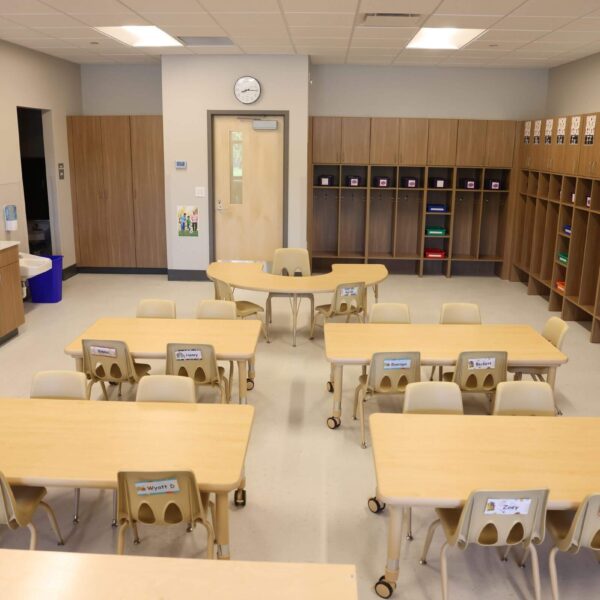 A classroom with light wooden tables and chairs arranged neatly. Shelves along the walls hold colorful bins. A clock and whiteboard are visible at the front. The room is well-lit with a modern and organized appearance.