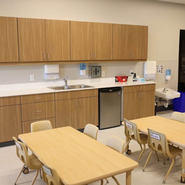 A classroom kitchenette with wooden cabinets, a sink, and a small fridge. Two tables with beige chairs are in the foreground. Soap dispensers and various supplies are on the countertop. A door is partially open, leading to another room.