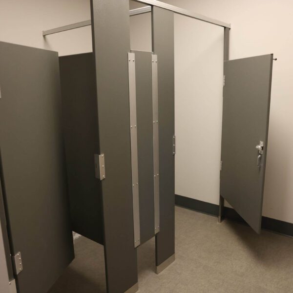 A public restroom stall with three gray partitions, two of which are closed and one slightly open, revealing an empty interior. The floor is tiled and the walls are plain white.