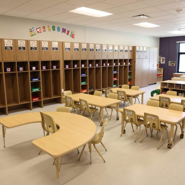 A tidy classroom with light wooden tables and chairs arranged in groups. Colorful storage cubbies line the back wall with welcome decorations above. Bright, natural light enters through large windows, creating a cheerful atmosphere.